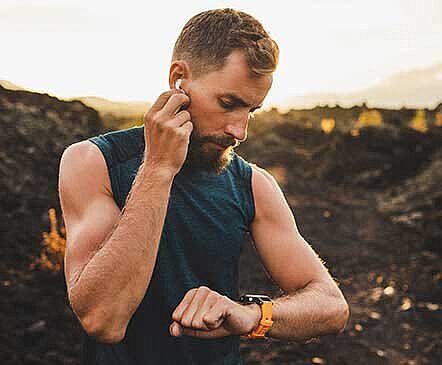 Bearded man in dark sleeveless shirt adjusts earbud while checking orange sports watch outdoors at sunset