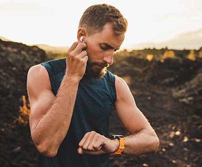 Bearded man in dark sleeveless shirt adjusts earbud while checking orange sports watch outdoors at sunset
