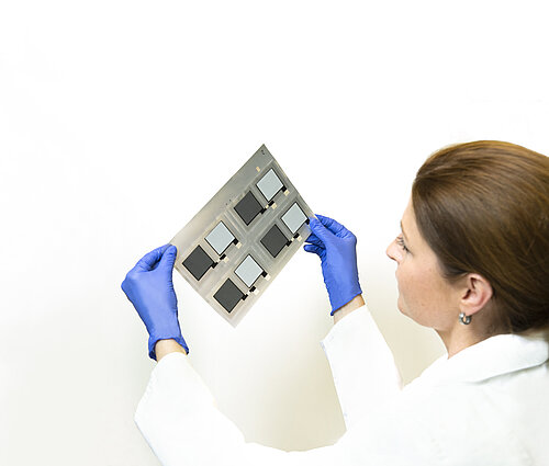 Researcher in white lab coat and blue gloves examines a substrate with six square thin-film samples against white background