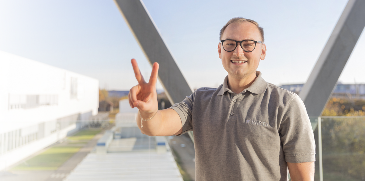 Smiling man with glasses in grey VARTA polo shirt making peace sign, standing on building walkway in sunlight