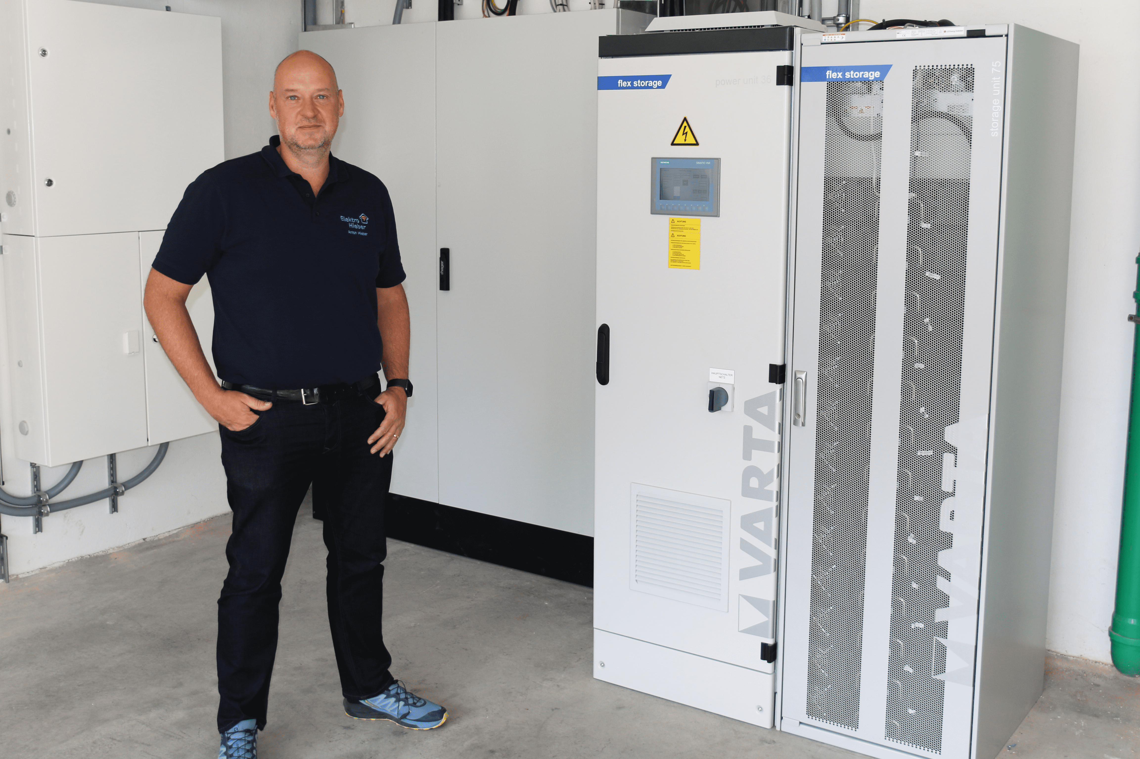 Mann in dunklem Poloshirt steht neben zwei VARTA-Batteriespeicherschränken mit Beschriftung flex storage in einem Technikraum
