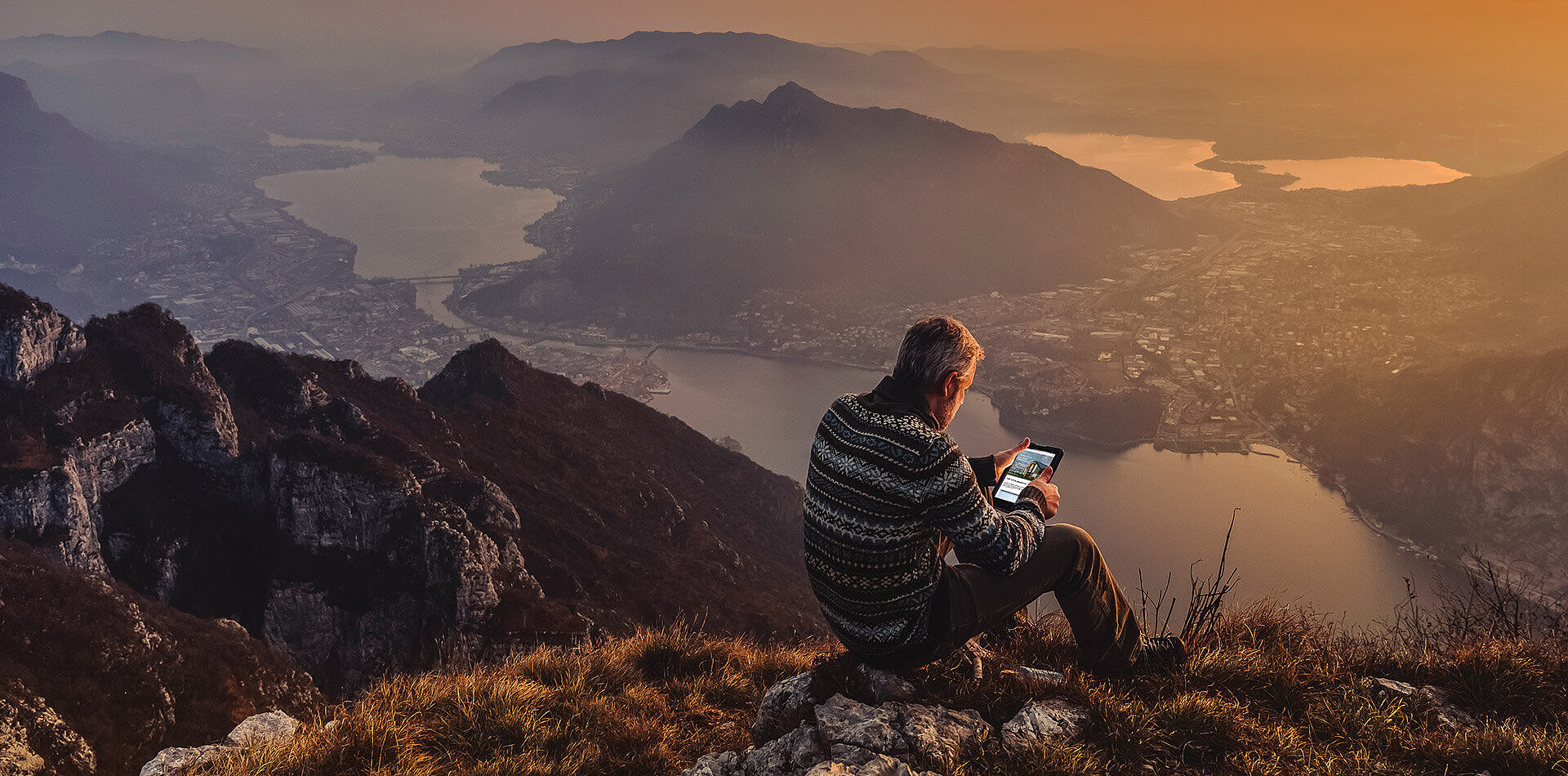 Man in patterned sweater sits on rocky mountain peak using tablet, overlooking lake, valley and hazy mountains at sunset