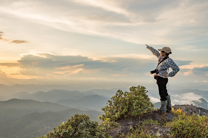 Persona con gorra y cámara señala el horizonte desde una cima rocosa con vistas a montañas brumosas al atardecer