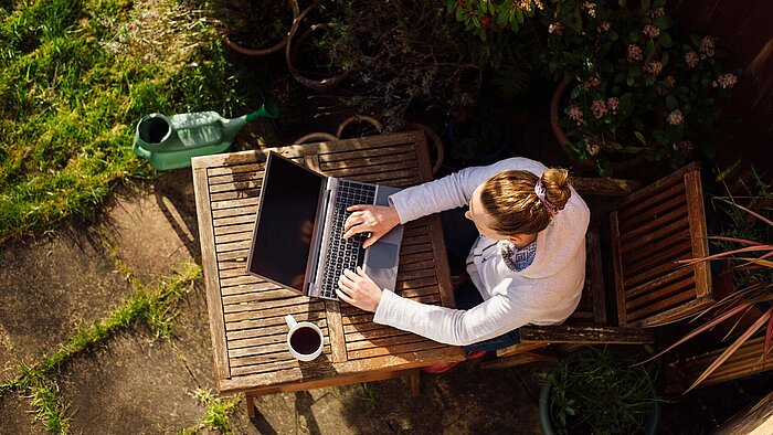 Overhead view of woman working on laptop at wooden garden table with coffee mug, watering can and potted plants nearby