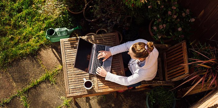 Overhead view of woman working on laptop at wooden garden table with coffee mug, watering can and potted plants nearby