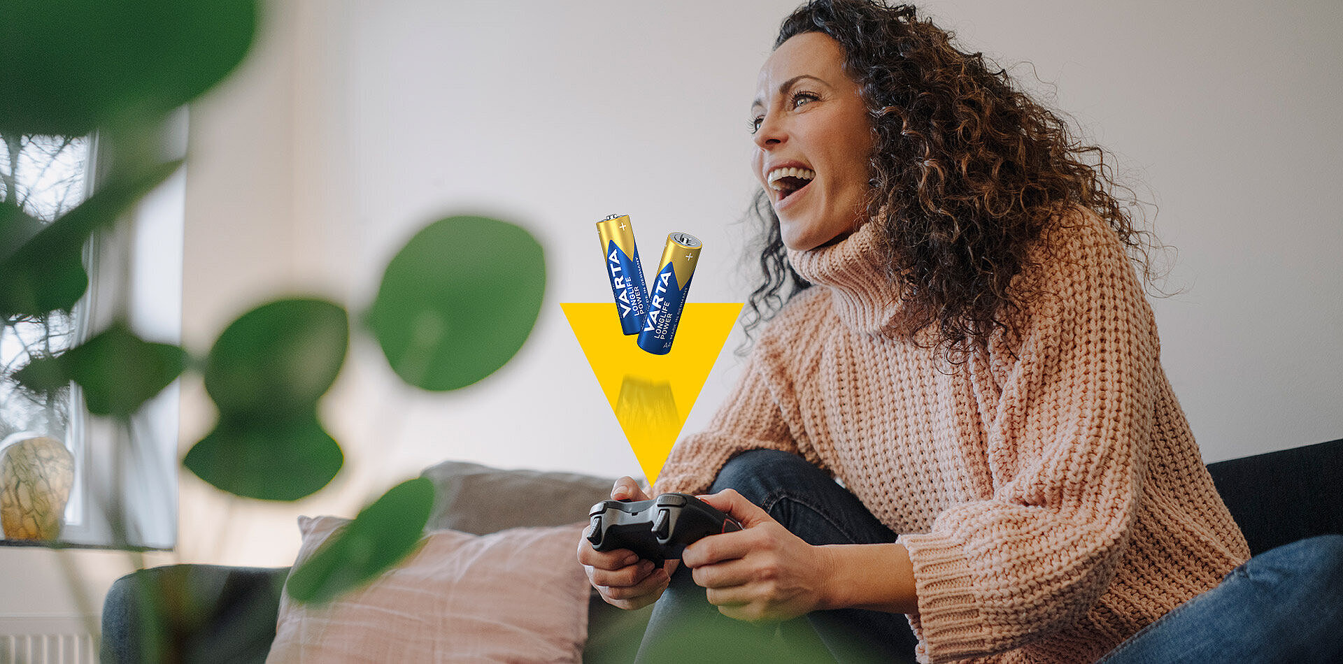 Laughing woman in beige sweater plays video game with controller, Varta Longlife Power batteries shown on yellow arrow