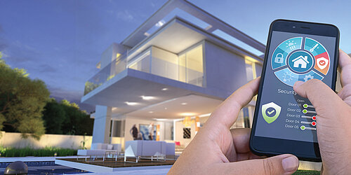 Hands holding a smartphone displaying a smart home security app in front of a modern illuminated house at dusk