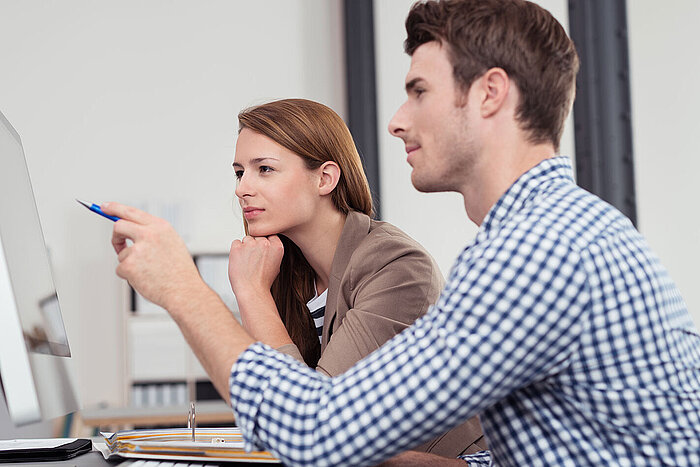 Two colleagues looking at a computer monitor, man in checkered shirt and woman in beige jacket pointing with a pen
