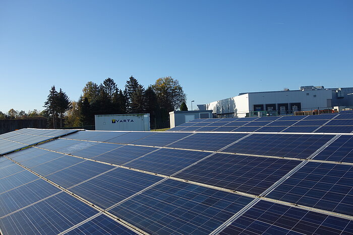 Große Photovoltaikanlage auf Flachdach, im Hintergrund Industriegebäude und VARTA-Container unter blauem Himmel