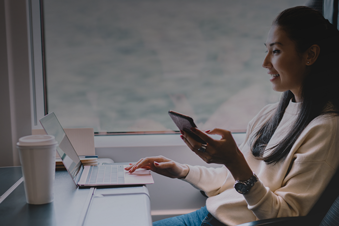 Smiling woman in cream sweater holds smartphone while using laptop at a table by a large window
