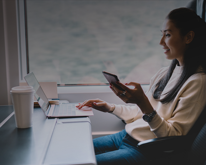 Smiling woman in cream sweater holds smartphone while using laptop at a table by a large window
