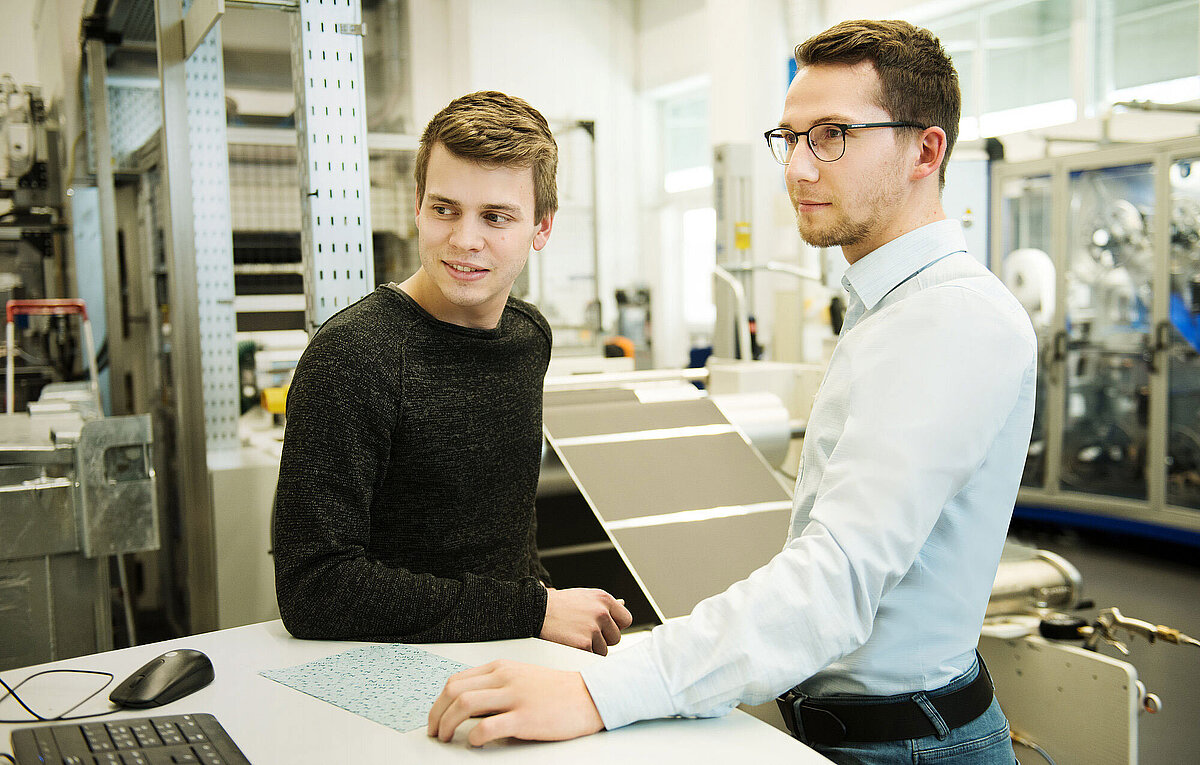 Two young men stand at a workbench in an industrial lab, one in a dark sweater, the other in a light shirt with glasses