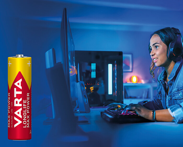 Junge Frau mit Headset spielt am Computer, links im Vordergrund eine Varta Longlife Max Power Batterie