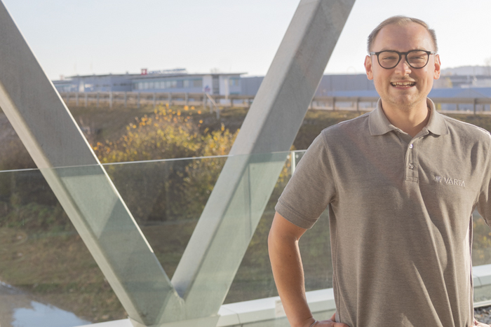 Lächelnder Mann mit Brille im grauen Varta-Poloshirt steht auf einer Brücke vor unscharfem Industriegebäude