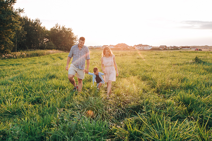 Familie mit Kind läuft bei Sonnenuntergang durch hohe Wiese, im Hintergrund Bäume und Häuser