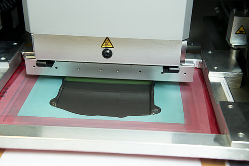 Screen printing machine with squeegee spreading black paste across a turquoise screen framed in red, with warning label visible