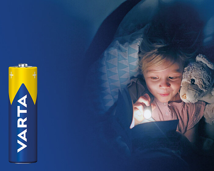 Child with stuffed bunny reading under blanket by flashlight, next to a Varta battery on blue background