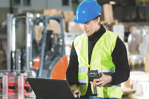 Warehouse worker in blue hard hat and yellow safety vest using barcode scanner and laptop near forklift