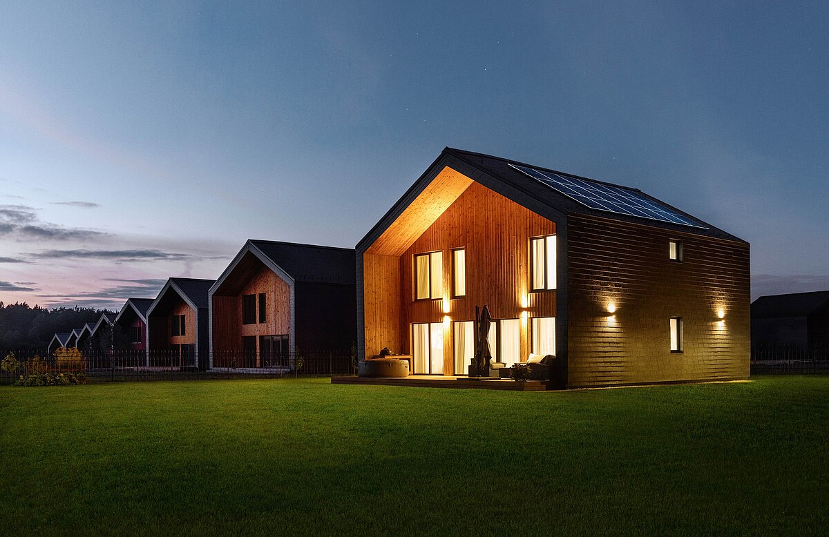 Modern wooden gable-roof houses with solar panels and lit interiors at dusk, set on a green lawn under a blue sky