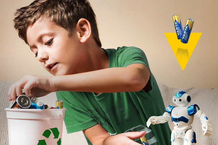 Bambino in maglietta verde mette batterie usate in un contenitore con simbolo del riciclo, accanto un robot giocattolo