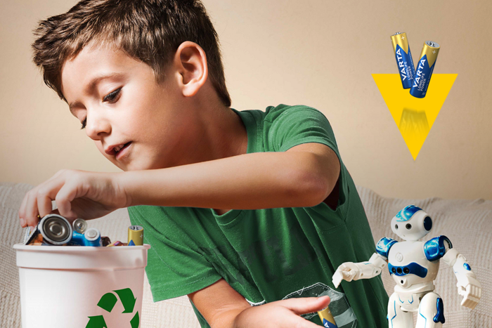Yeşil tişörtlü çocuk geri dönüşüm kutusuna piller atıyor, yanında oyuncak robot ve sarı ok ile Varta piller görseli