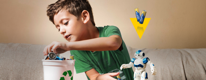 Boy in green shirt places used batteries into white recycling bin, blue robot toy beside him, Varta batteries above