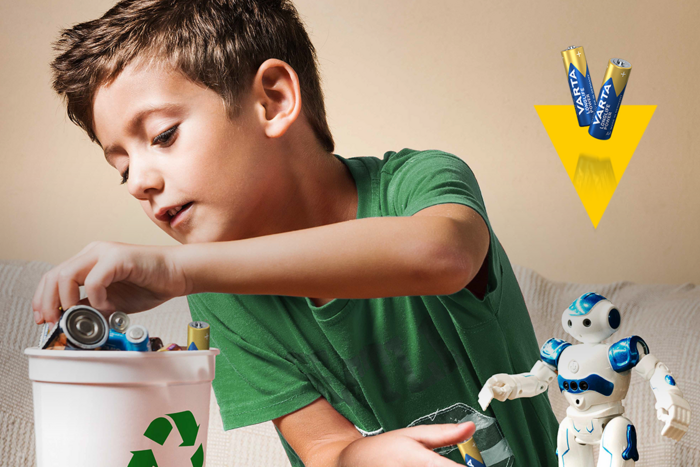 Junge in grünem T-Shirt wirft Batterien in Recyclingbehälter, daneben blauer Spielzeugroboter und Varta-Batterien