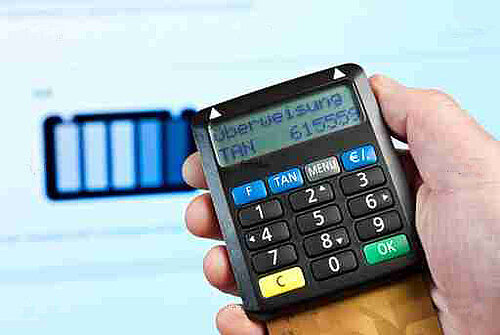 Hand holding a TAN generator device displaying "Überweisung" and a six-digit code on its screen