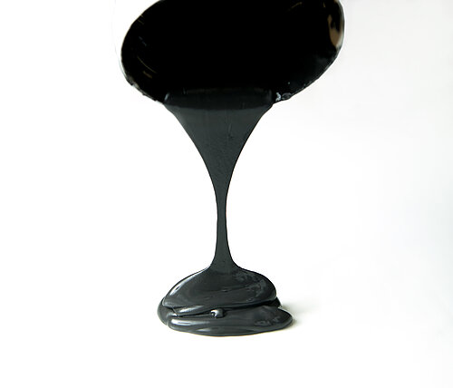 Thick black viscous liquid pouring downward in a continuous stream, forming a glossy pile on a white surface