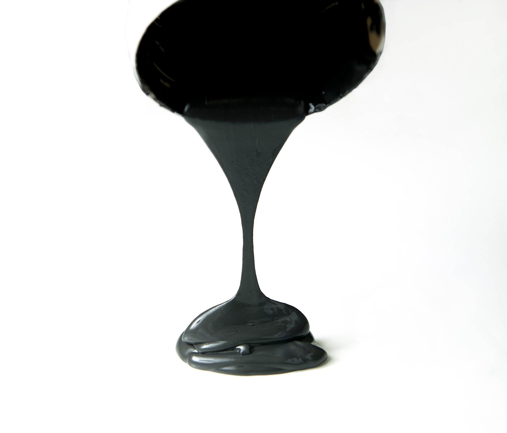Thick black viscous liquid pouring downward in a continuous stream, forming a glossy pile on a white surface