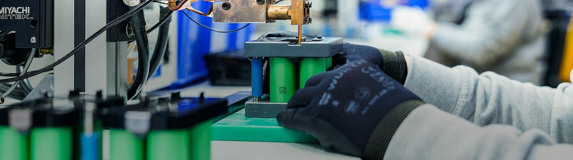 Gloved hand positioning green cylindrical battery cells under a copper welding tool in an industrial assembly setup