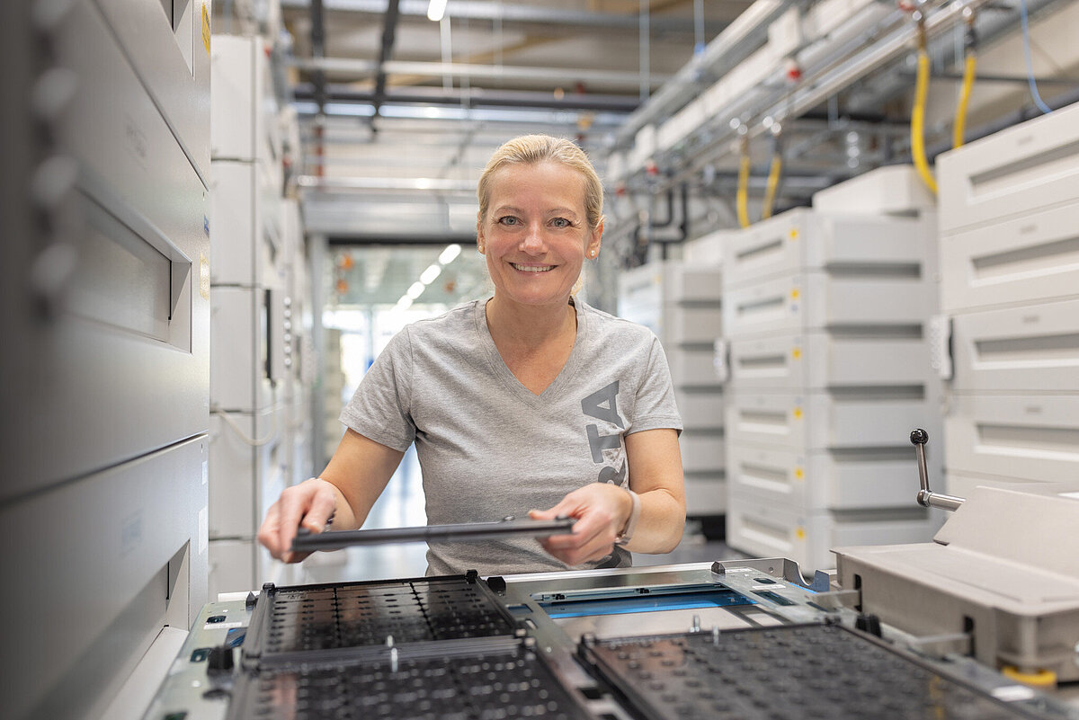 Smiling blonde woman in grey VARTA t-shirt working with equipment in industrial facility with white machinery cabinets
