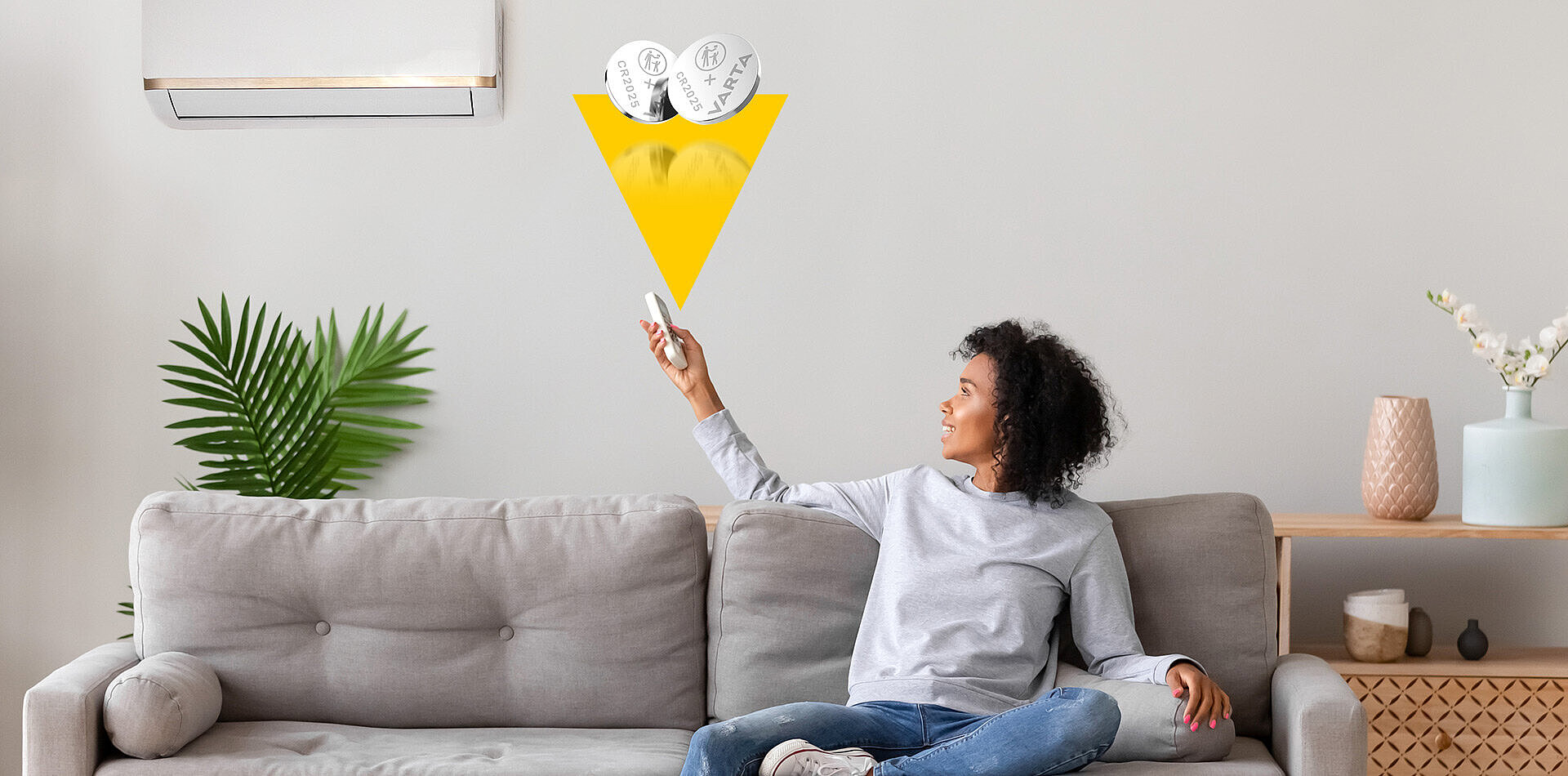 Smiling woman on grey sofa points remote at wall air conditioner, with two VARTA CR2025 batteries shown above in yellow arrow