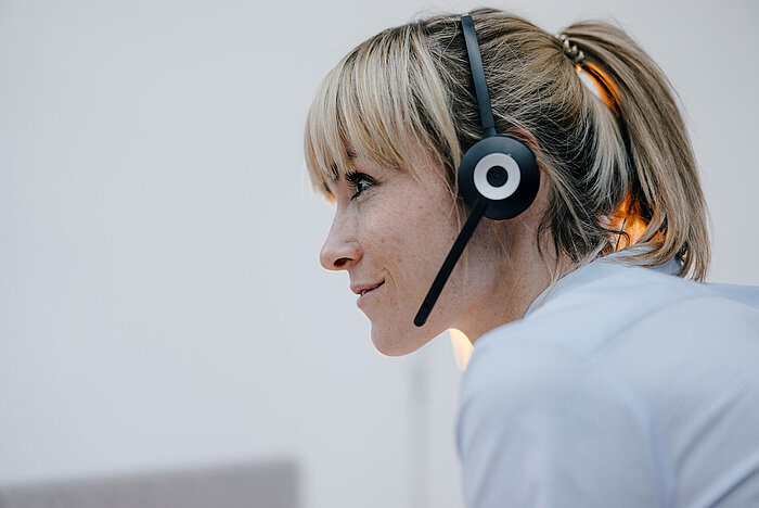 Blond kvinne med hestehale i profil, smiler og bruker svart headset med mikrofon, iført lyseblå skjorte