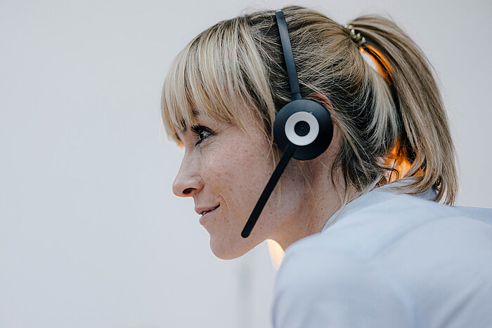 Blond kvinne med hestehale i profil, smiler og bruker svart headset med mikrofon, iført lyseblå skjorte