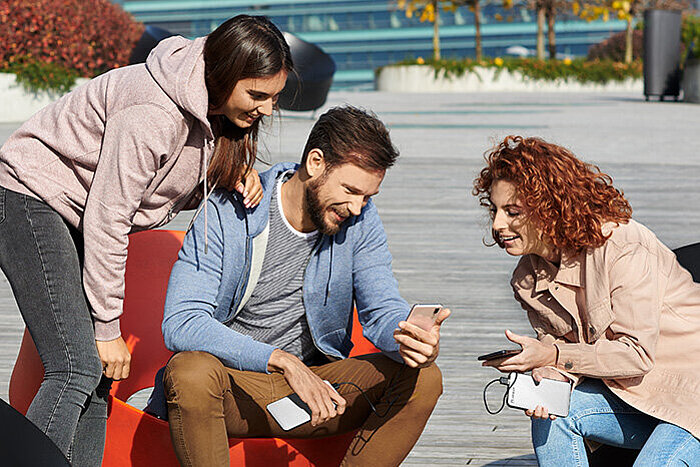 Three young friends sitting outdoors on orange seats, smiling while looking at smartphones together on a sunny day