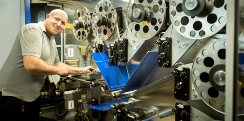 Smiling man in grey polo shirt operating an industrial film reel machine with large metal reels and blue components