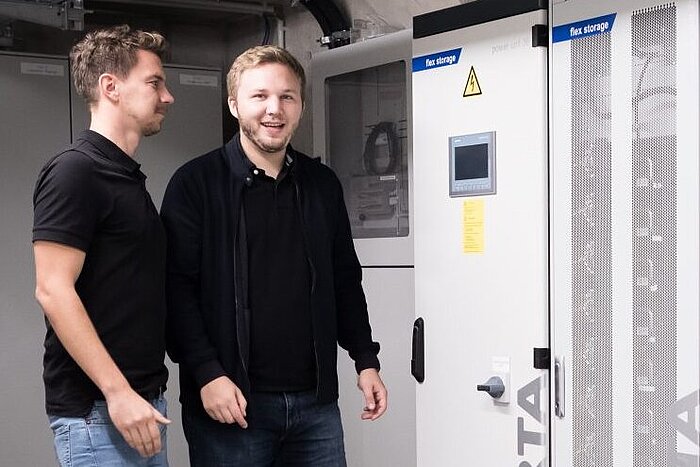 Zwei junge Männer in dunkler Kleidung stehen vor einem Batteriespeicherschrank mit Bedienpanel und Beschriftung flex storage
