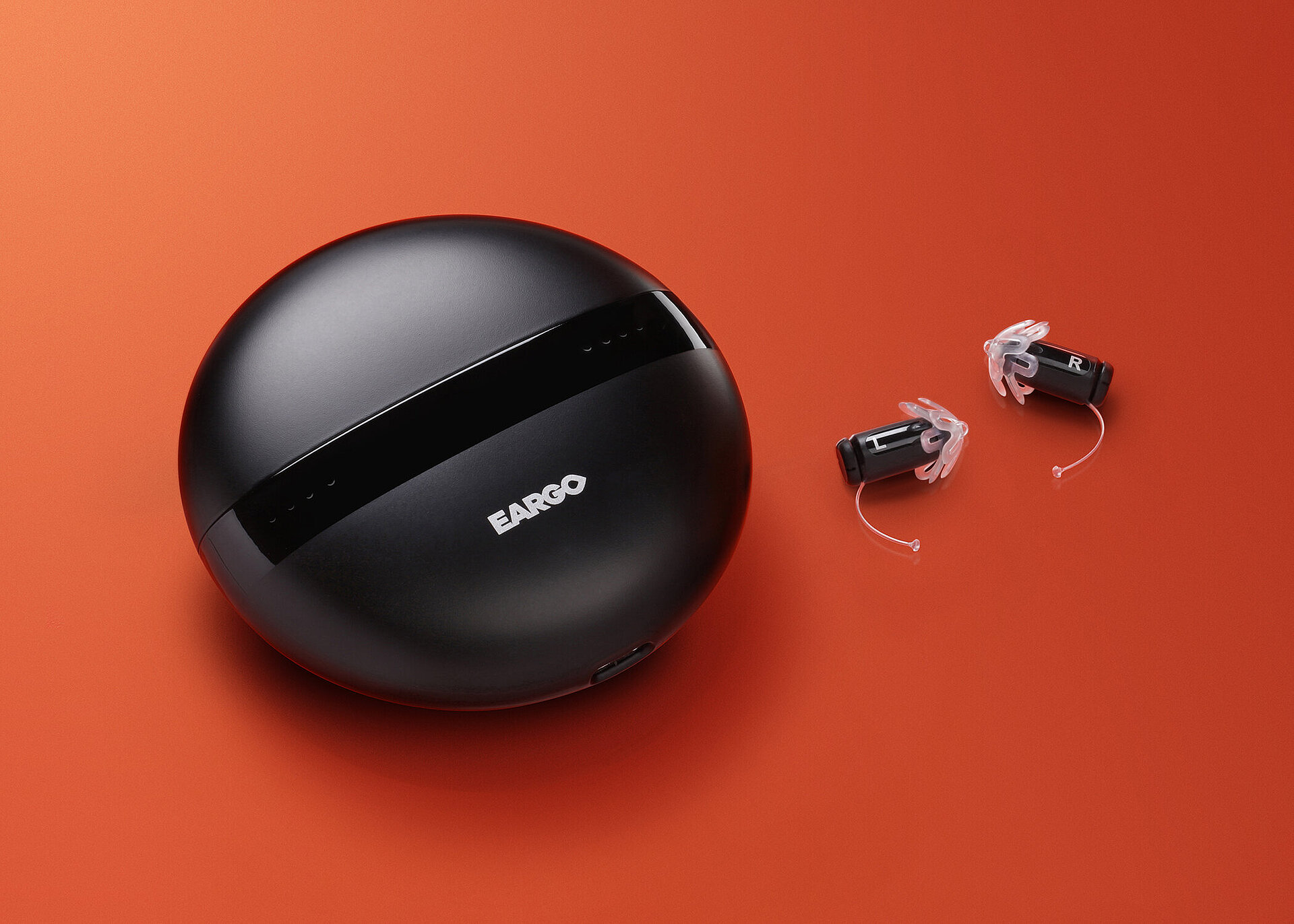 Round black Eargo charging case with two small hearing aids labeled L and R on an orange background