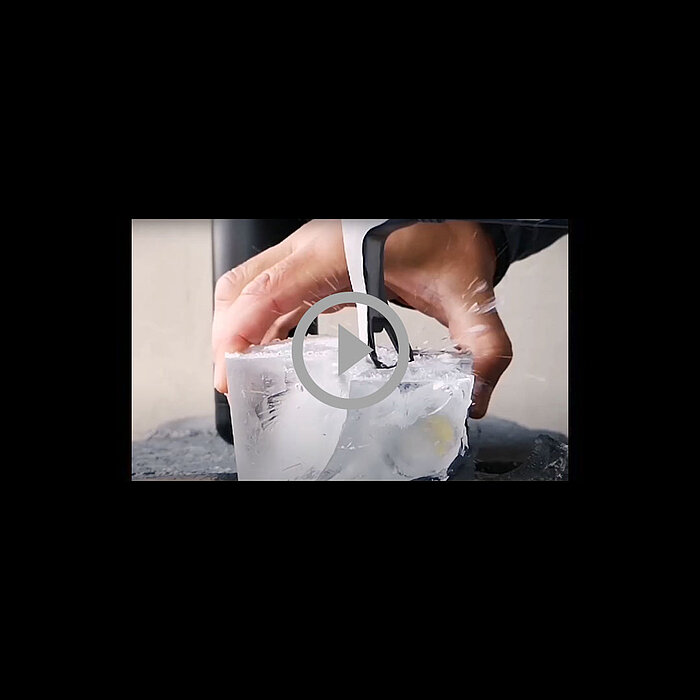 Hands shaping a large block of ice with a tool, video play button overlay in the center