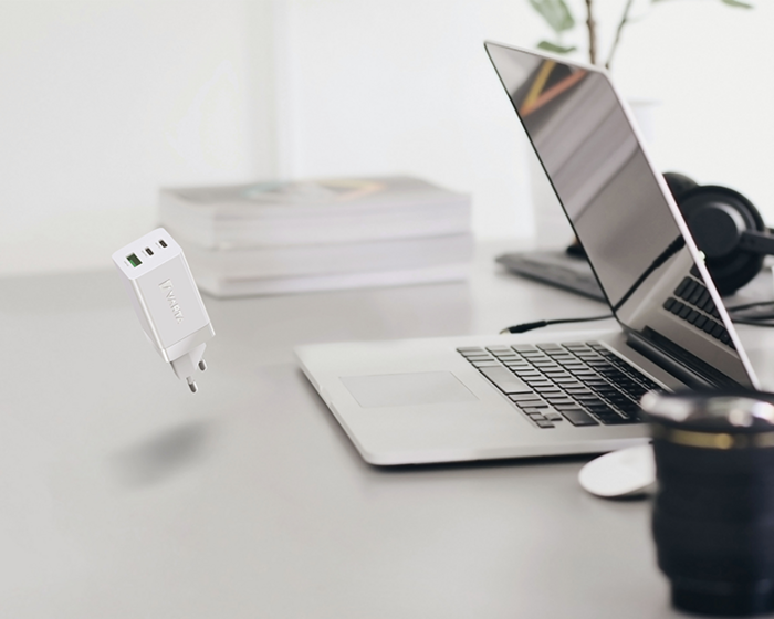 White compact USB-C power adapter floating above desk near open laptop, headphones, books and camera lens