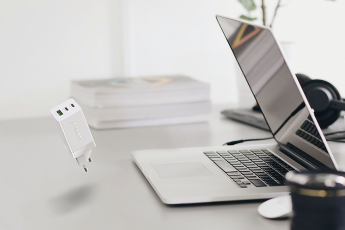 Gri masa üzerinde açık dizüstü bilgisayar, kulaklık, kitap yığını ve havada süzülen beyaz USB şarj adaptörü