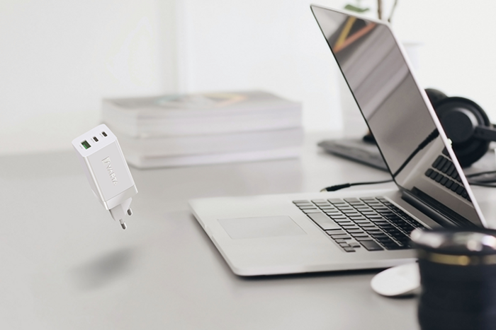 White compact USB-C power adapter floating above desk near open laptop, headphones, books and camera lens