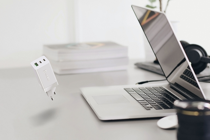 Caricatore USB bianco sospeso in aria sopra una scrivania con laptop aperto, libri, cuffie e obiettivo fotografico