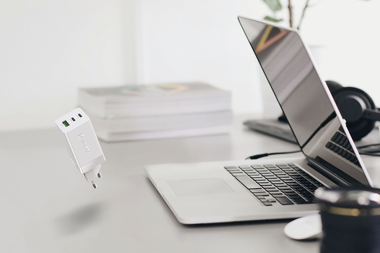 Valkoinen USB-laturi leijuu työpöydän yllä, taustalla avoin kannettava tietokone, kuulokkeet, kirjapino ja kameran objektiivi