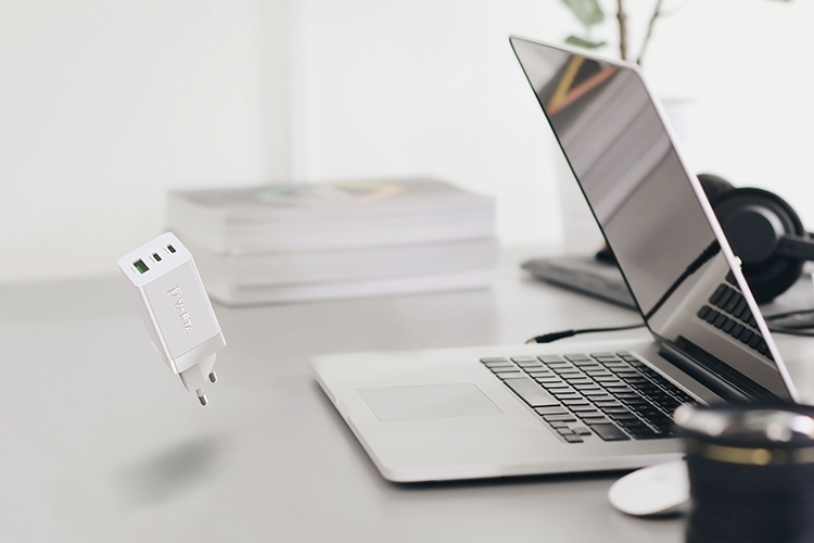 Gri masa üzerinde açık dizüstü bilgisayar, kulaklık, kitap yığını ve havada süzülen beyaz USB şarj adaptörü