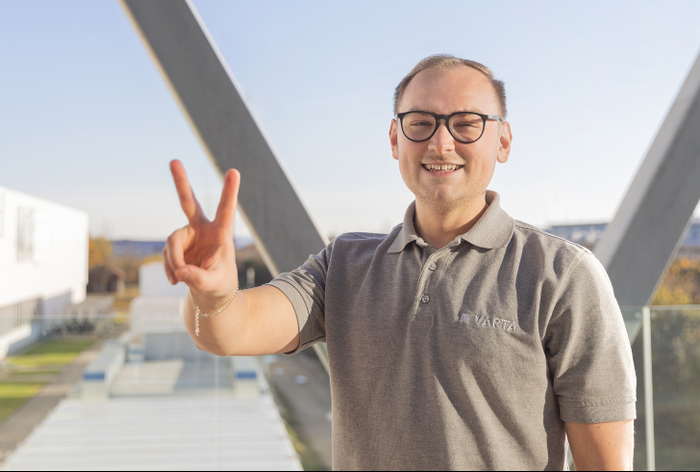 Smiling man with glasses in gray Varta polo shirt making peace sign, standing outdoors near modern building