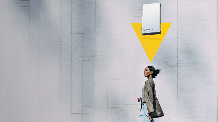 Woman with rolling suitcase walking past concrete wall with mounted VARTA battery unit and yellow downward arrow