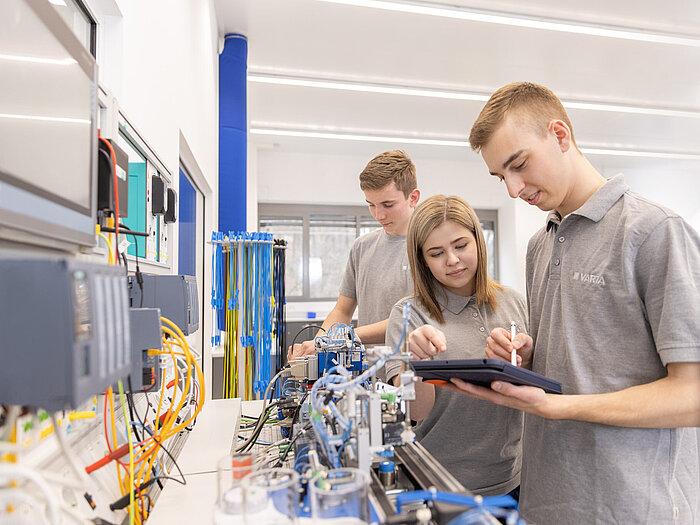 Drei junge Personen in grauen Poloshirts arbeiten an technischer Anlage, eine hält ein Tablet, im Hintergrund Steuerungstechnik
