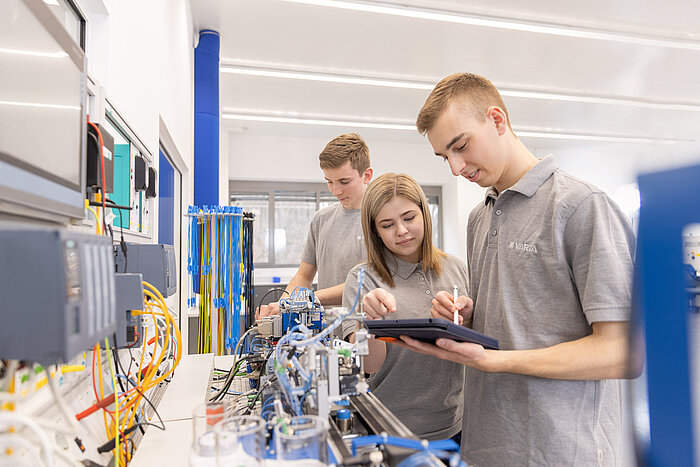 Three young technicians in grey polo shirts examine a tablet while working on industrial automation equipment in a lab